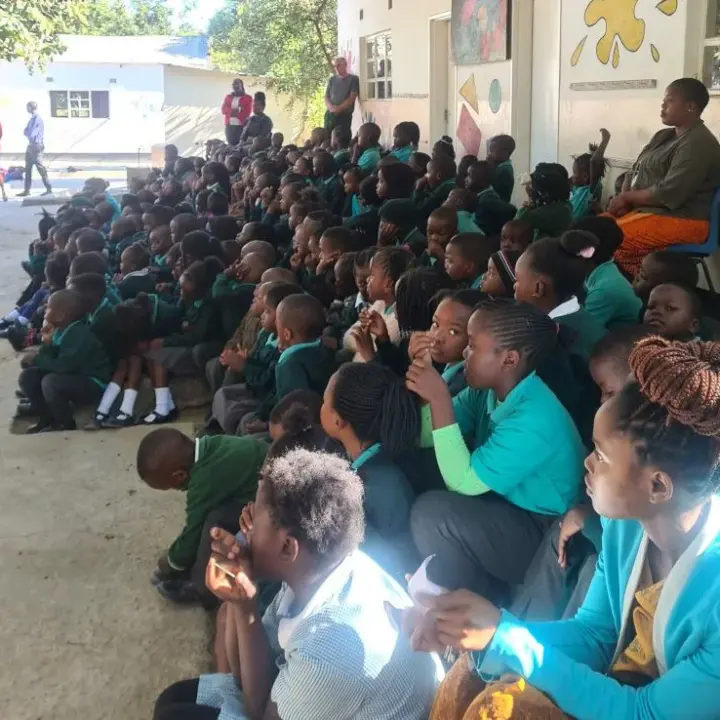 Large group of schoolchildren in uniform seated outdoors facing right, with adults standing and sitting nearby
