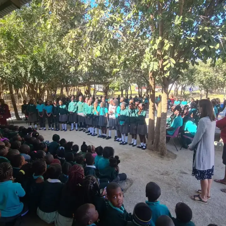 Group of schoolchildren in green uniforms gathered outdoors under trees with a line of students standing and others seated