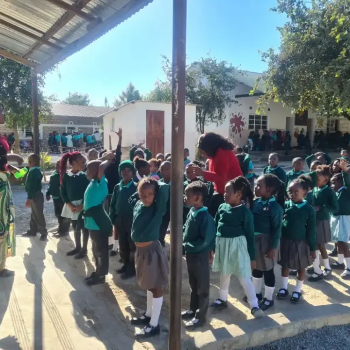 Group of young schoolchildren in green uniforms lined up outdoors with two adult teachers under a roofed area