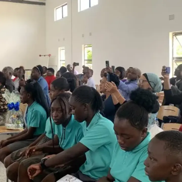 Students in teal uniforms seated in rows with adults behind them filming on phones in a bright classroom