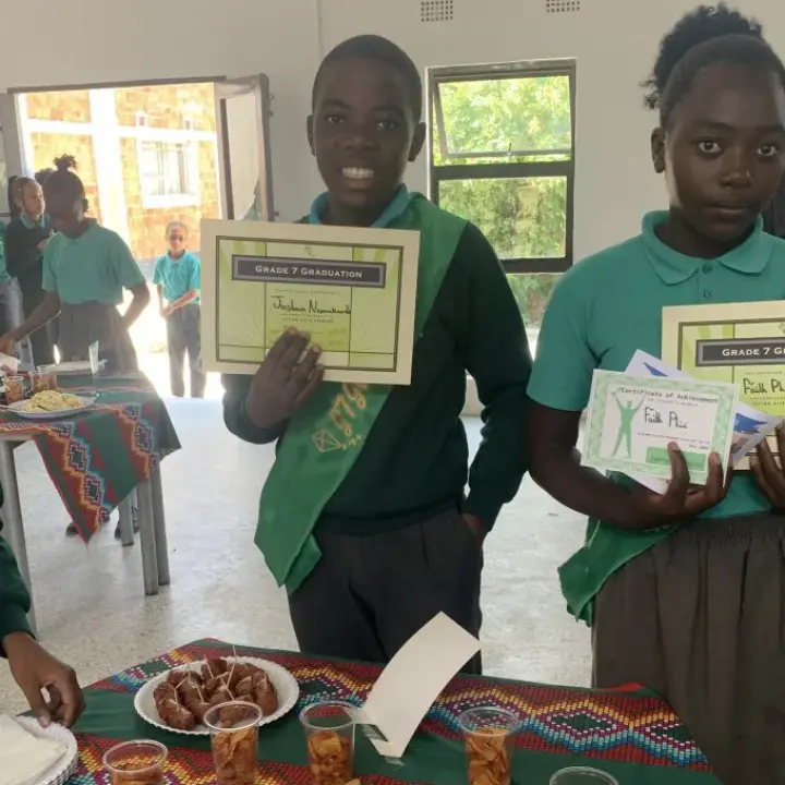 Two students wearing green sashes holding graduation certificates standing behind a table with snacks and cups