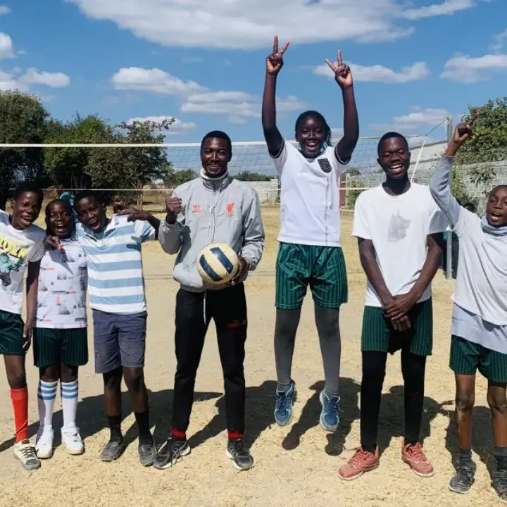 Seven smiling children and a coach standing on a dusty volleyball court, one child jumping with arms raised and the coach