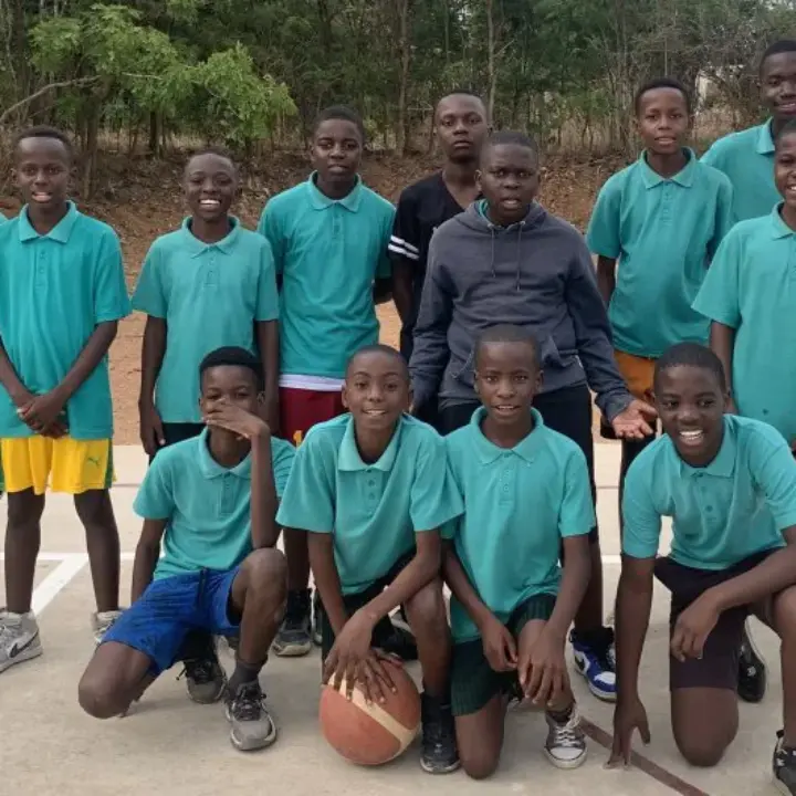 Group of thirteen boys on an outdoor basketball court, most wearing teal polo shirts; one holds a basketball.